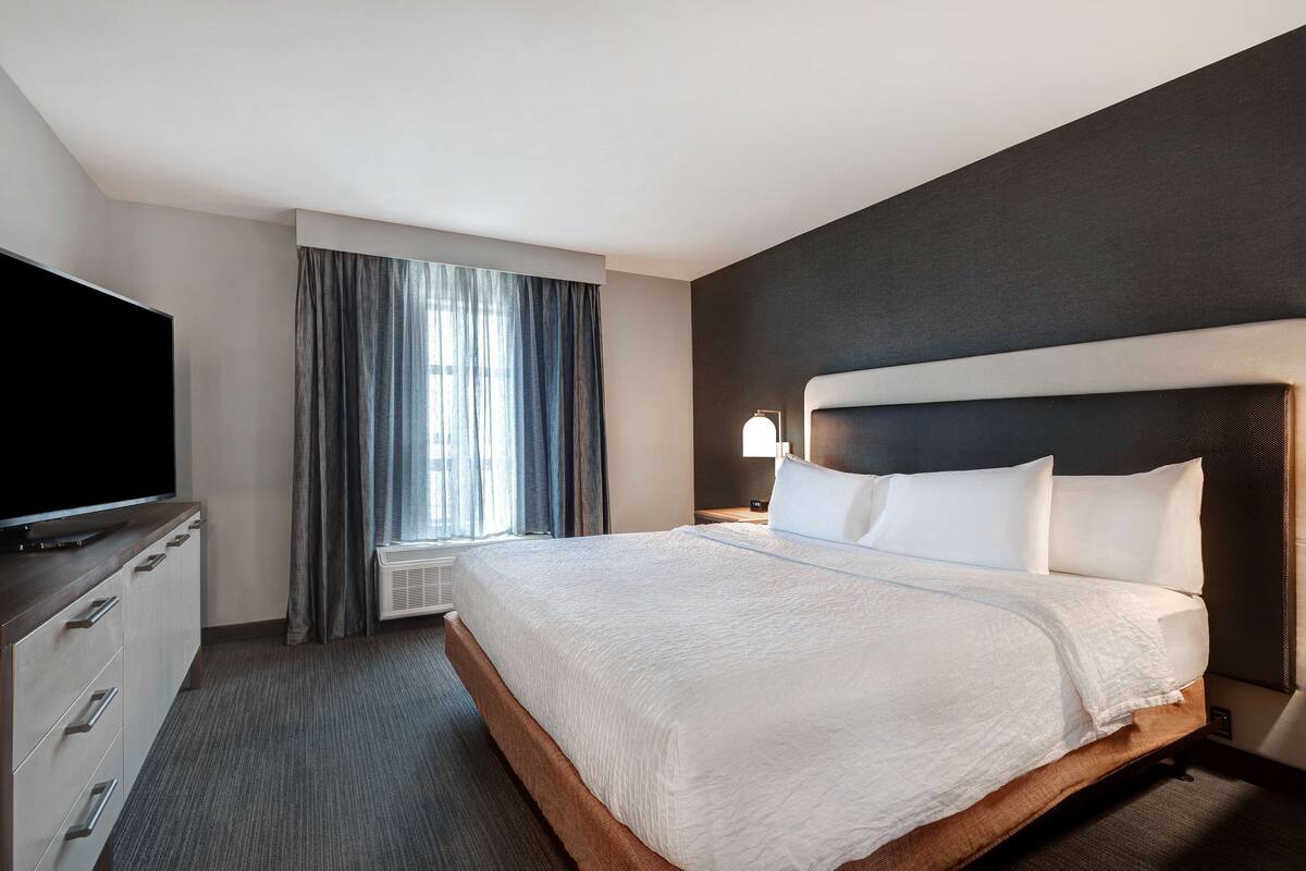 Habitación de hotel con una cama grande, un televisor de pantalla plana, un aparador y una ventana con cortinas.
