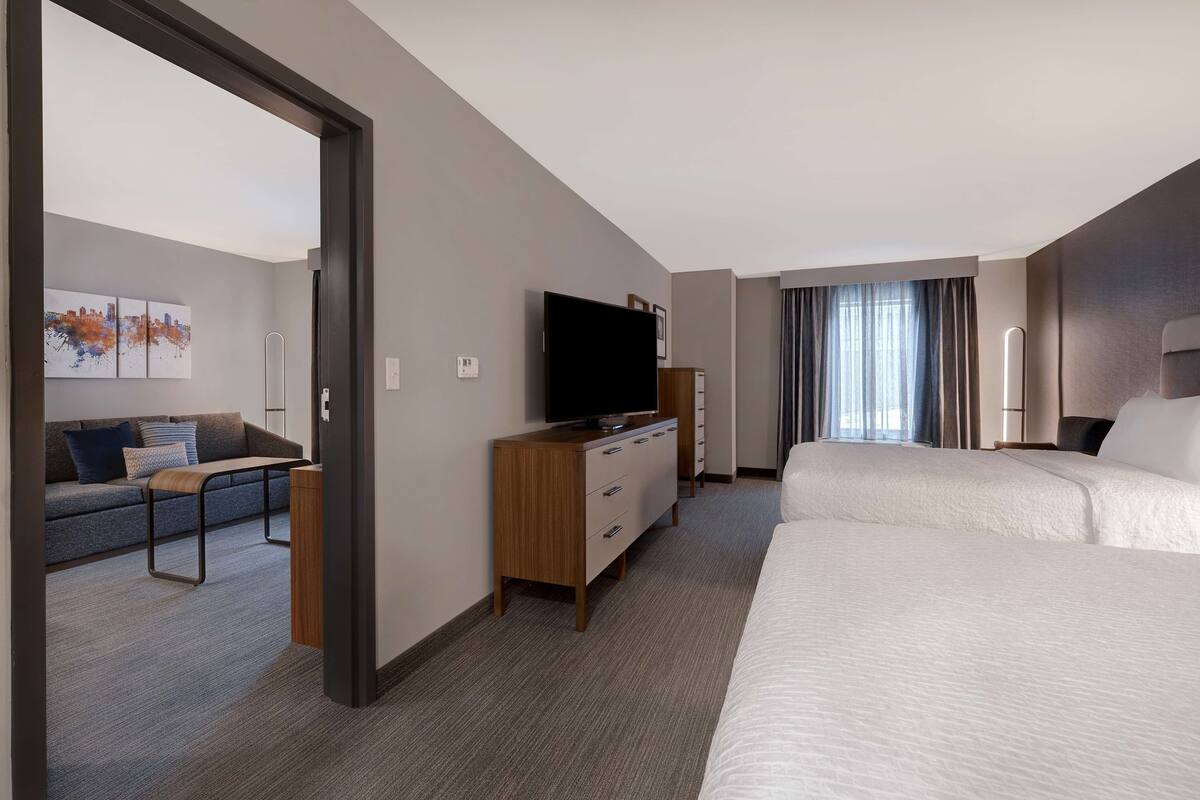 Una habitación de hotel con una cama grande, un sofá, un televisor sobre un soporte de madera y una obra de arte con vistas a la ciudad.