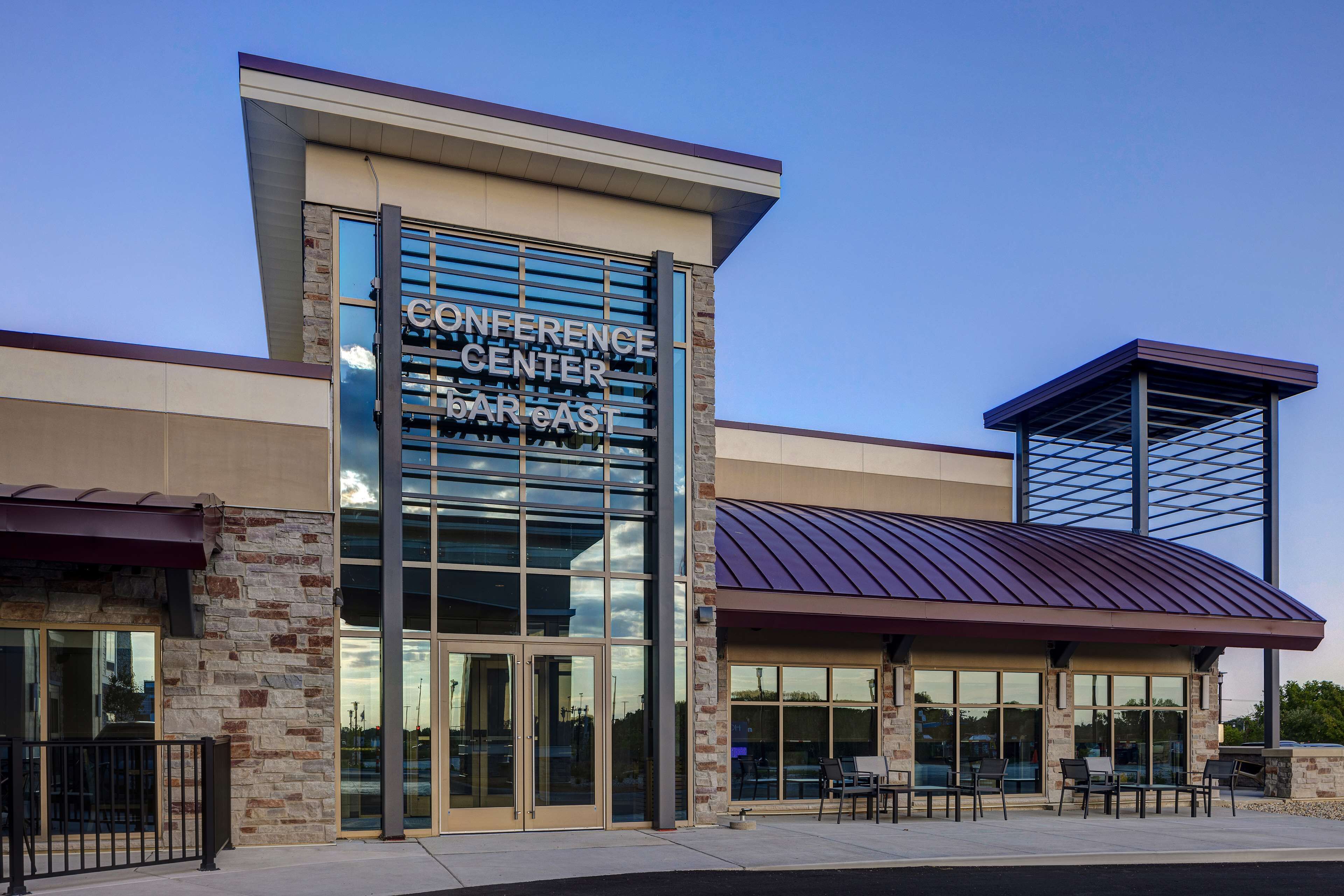 Un edificio moderno con un letrero que dice "Conference Center Kansas City". Cuenta con amplias ventanas de vidrio, fachada de ladrillo y una zona de estar exterior cubierta.