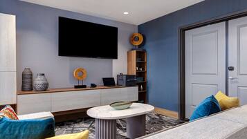 A modern living room with a flat-screen TV mounted on the wall, a wooden console table with decorative items, a round coffee table, and a sofa with colorful cushions.