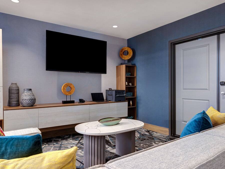 A modern living room with a flat-screen TV mounted on the wall, a wooden console table with decorative items, a round coffee table, and a sofa with colorful cushions.