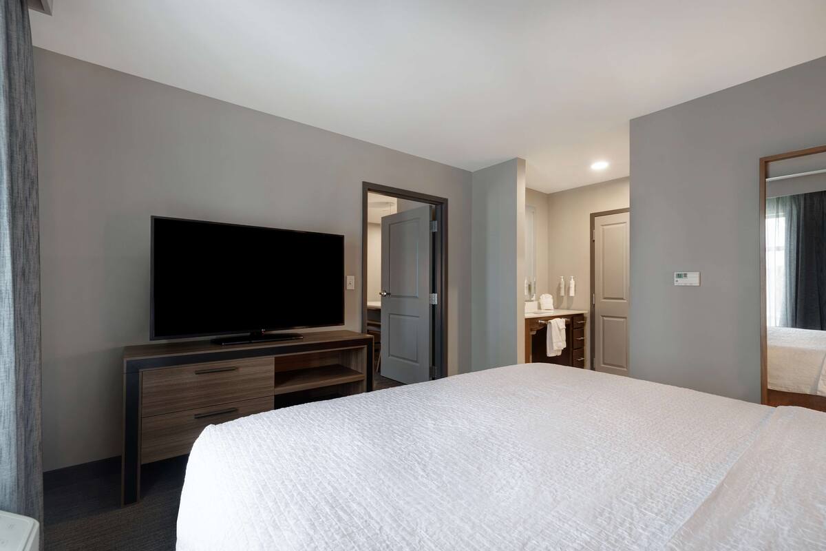 Habitación de hotel con una cama grande, un televisor sobre un mueble de madera, un baño con espejo y una puerta que da a otra habitación.