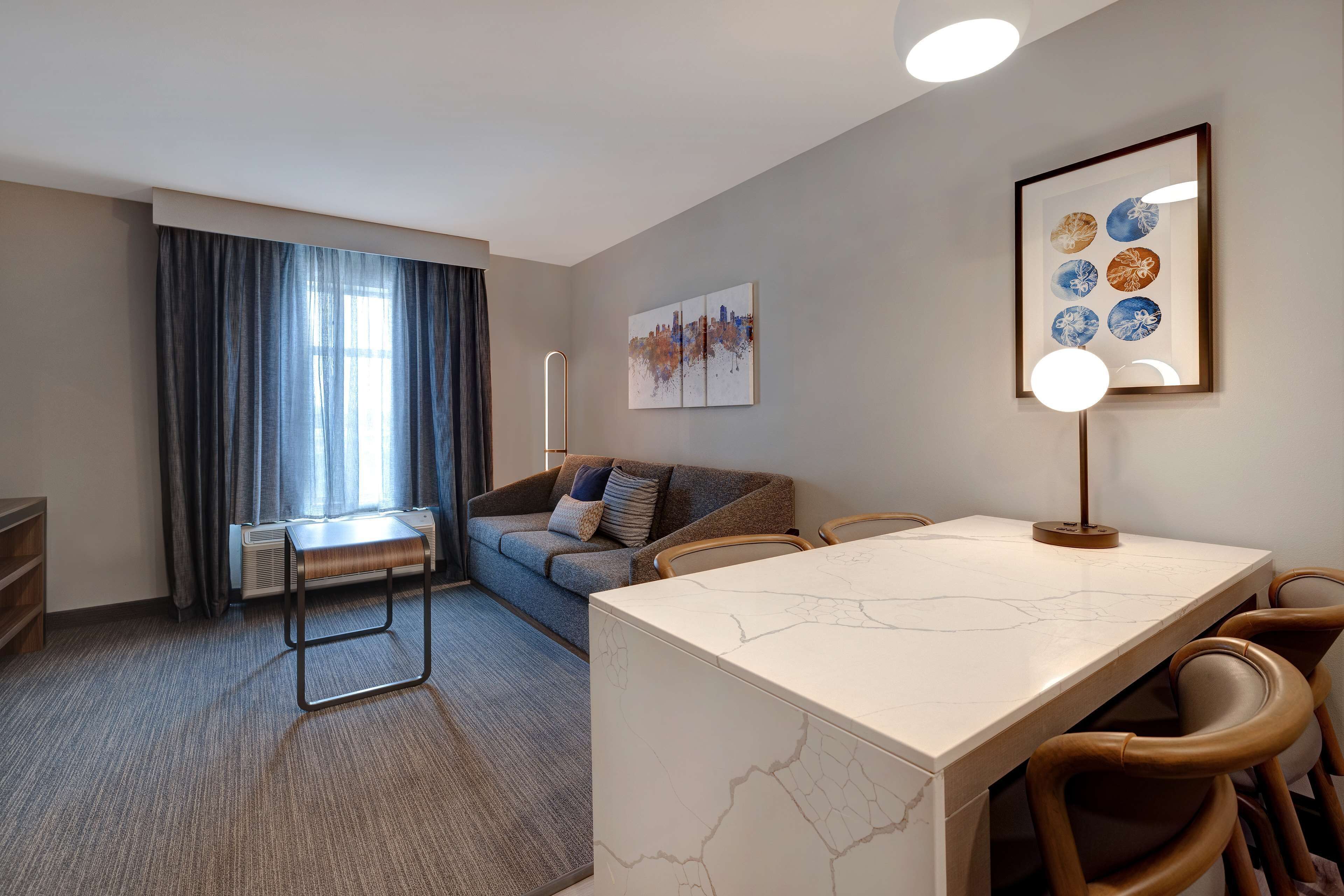 Una habitación de hotel moderna con una mesa de comedor de mármol, un sofá, una lámpara de pie y un gran ventanal con cortinas.