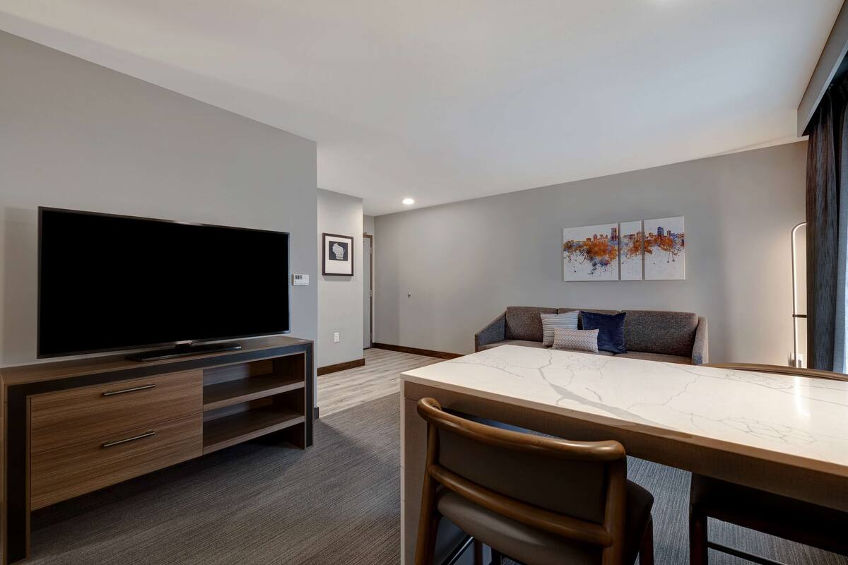 Una habitación de hotel moderna con un televisor de pantalla plana, una mesa de comedor con sillas, un sofá y una obra de arte con un paisaje urbano en la pared.