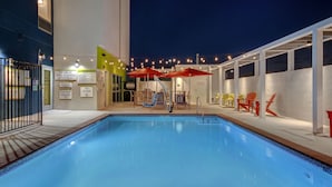 Une piscine dont le bord est revêtu de carrelage, entourée d’un espace de détente extérieur et illuminée de guirlandes lumineuses.