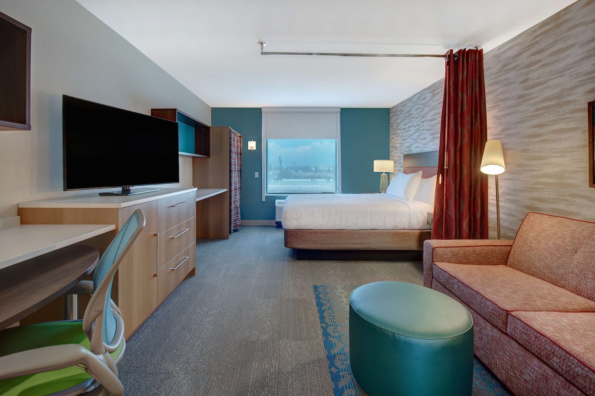 Une chambre d’hôtel avec un lit, un bureau avec une télévision, un canapé et une fenêtre avec des rideaux.