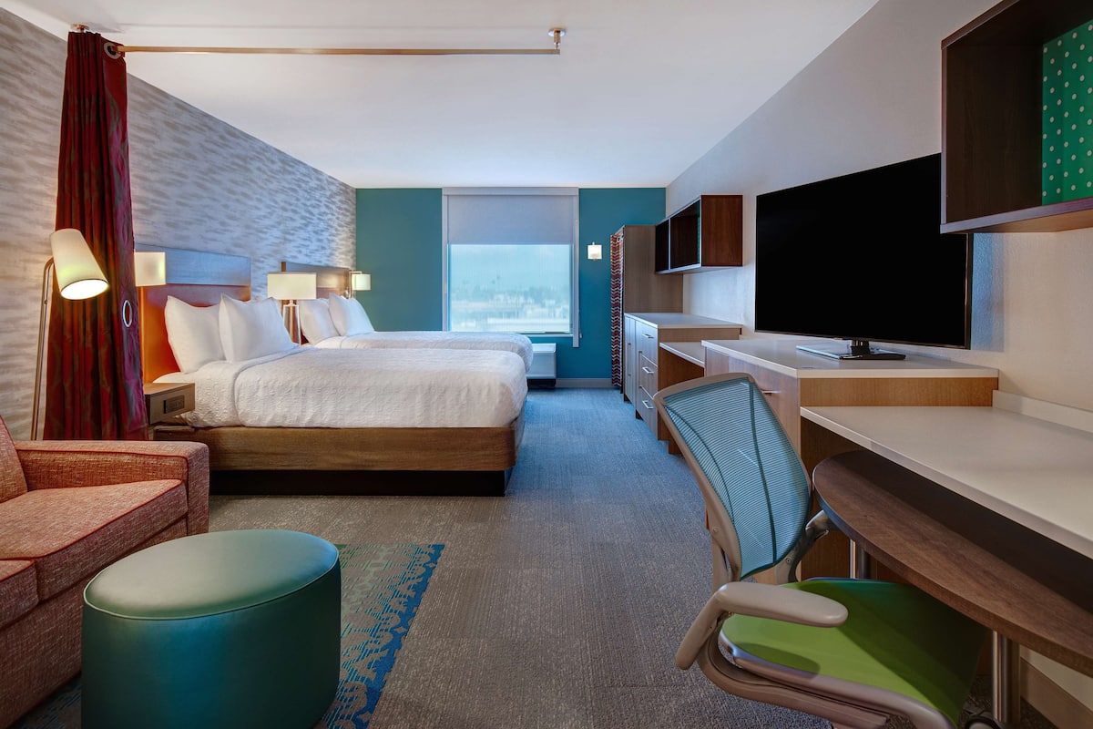 Une chambre d’hôtel comprenant un lit, deux tables de chevet, un bureau avec une chaise, une télévision et une fenêtre.