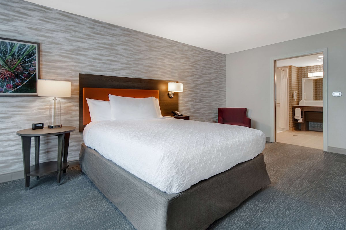 Une chambre d’hôtel moderne avec un grand lit, une table de chevet avec une lampe, un fauteuil rouge et une salle de bain visible à travers une porte ouverte.