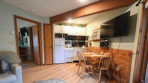 Una cocina compacta con electrodomésticos blancos, una zona de comedor con sillas de madera y un televisor montado en la pared.