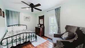 Ein Schlafzimmer mit Bett, Stuhl, Schminktisch, Deckenventilator und einem Fenster mit Vorhängen.