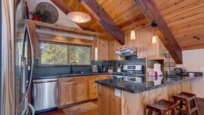 A modern kitchen with wooden cabinets, a stainless steel refrigerator, a large island with bar stools, and a window providing a view of the outdoors. - Edelweiss - Remodeled Luxury 3 BR with Private Hot tub (Tahoe City)