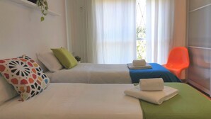 Een hotelkamer met twee bedden, een groene en een blauwe spreidek, wit beddengoed, een kleurrijk kussen, een kleine plant op een plank en een raam met gordijnen.