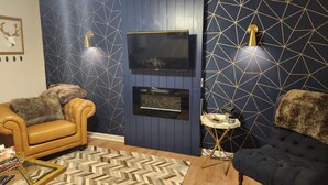 Un soggiorno moderno con una parete azzurra a motivi geometrici, una TV a schermo piatto montata, un divano in pelle, una poltrona scura e un tavolino con una lampada.