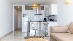 A modern kitchen with white cabinets, a stainless steel sink, and a bar stool. There is a beige sofa in the living area with a woven coffee table. - Nice apartment for 2 people with A/C, WIFI, TV, terrace and pets allowed (Empuriabrava)