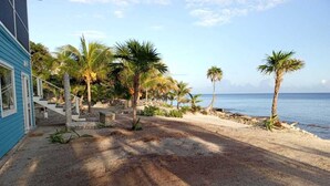 Il s’agit d’une propriété située en bord de mer, avec un sentier sablonneux menant à un bâtiment à la façade bleue. Des palmiers sont disséminés dans les environs.