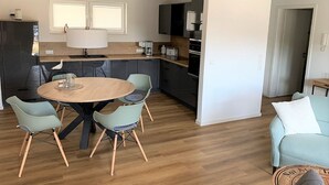 A modern kitchen with a round dining table, two chairs, a coffee machine, and a window with blinds. - 50.1.m Ankerplatz - Moderne, Hochwertige Ferienwohnung, Sauna, Großer Balkon, Strandnah (Wangerland)