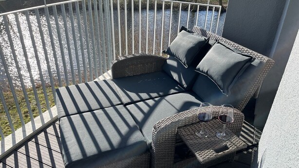 A wicker sofa with cushions on a balcony overlooking a body of water. - 50.1.m Ankerplatz - Moderne, Hochwertige Ferienwohnung, Sauna, Großer Balkon, Strandnah (Wangerland)