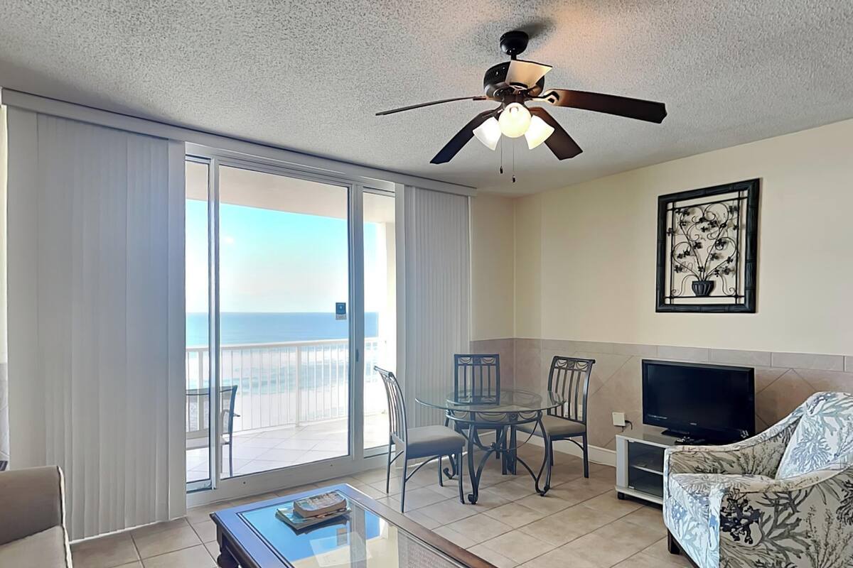 A living room with a glass coffee table, a sofa, a TV, and a balcony with a view of the ocean.