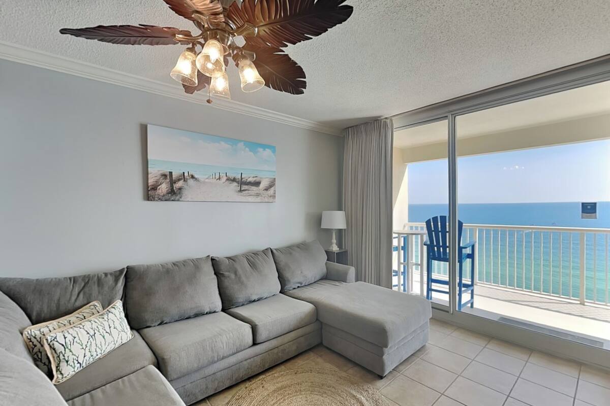 A living room with a grey sectional sofa, a round woven rug, a ceiling fan, a painting of a beach, and a balcony with a view of the ocean.
