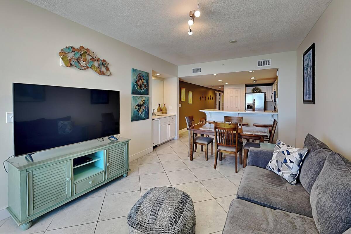 A living room with a flat-screen TV, a green entertainment unit, a gray sofa, and a dining area with a table and chairs.