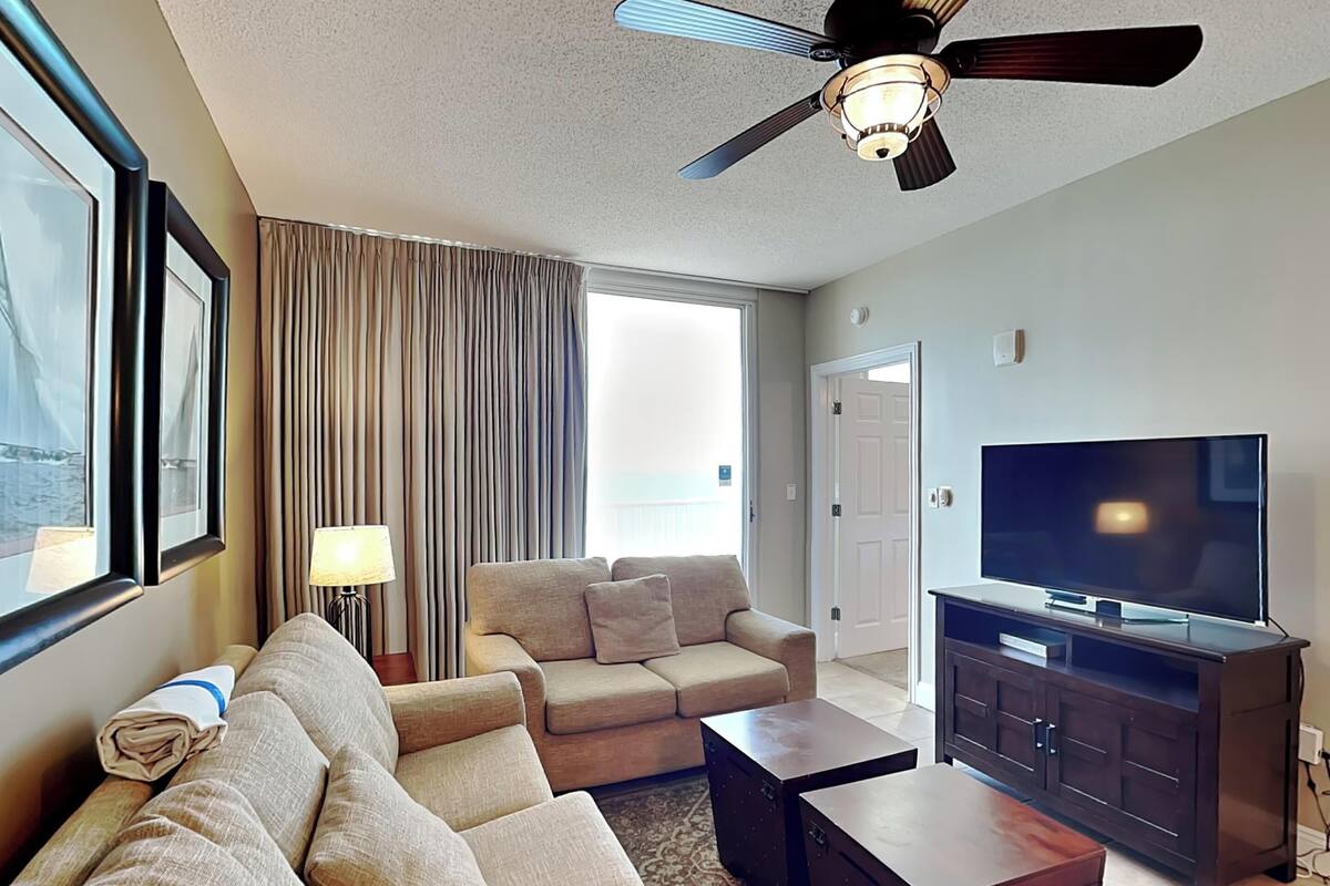 A hotel room with a large sofa, a television, a ceiling fan, and a view of the sea.