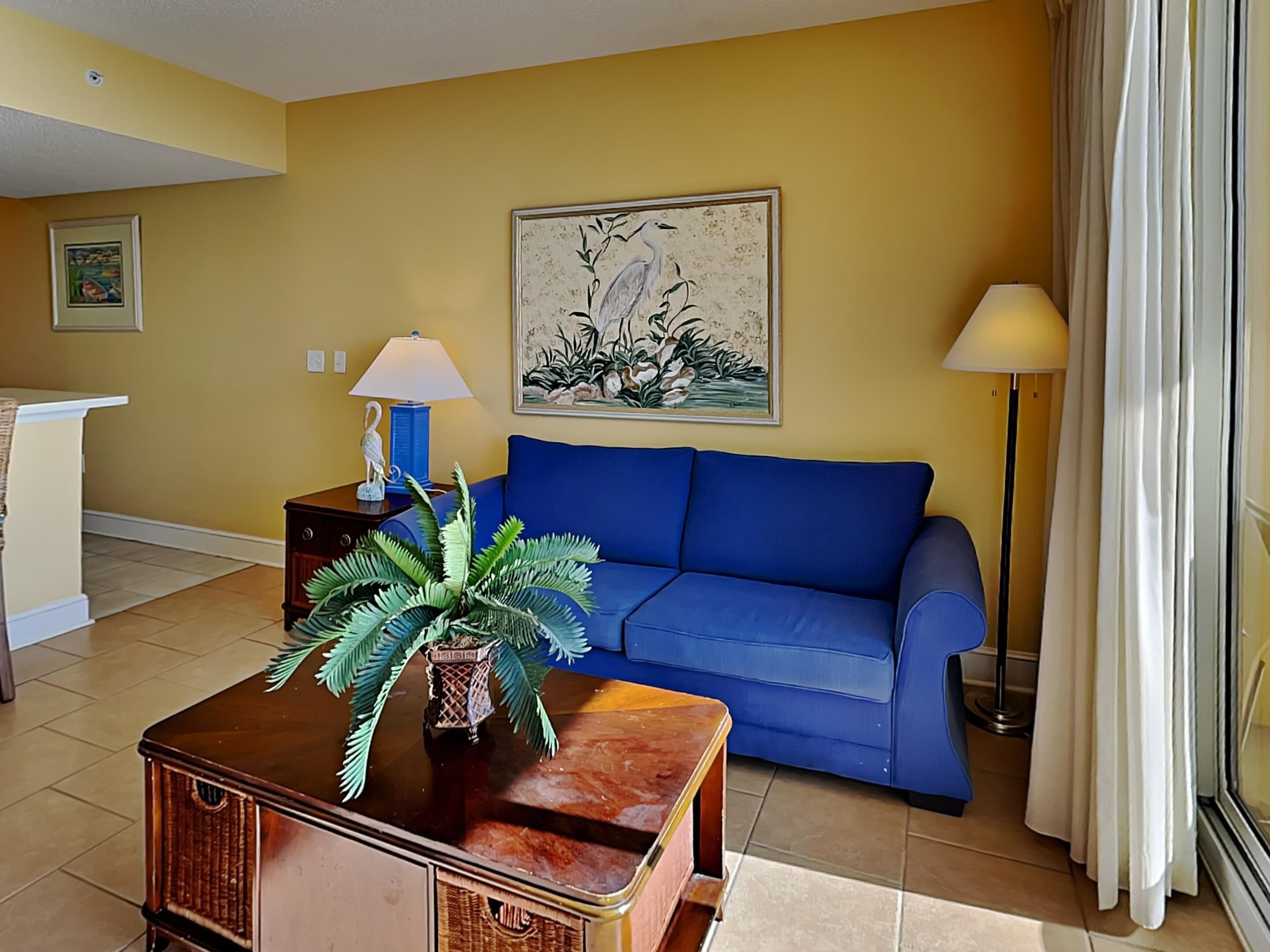 Uma sala de estar com um sofá azul, uma mesa de centro de madeira com uma planta, um quadro na parede e um abajur.