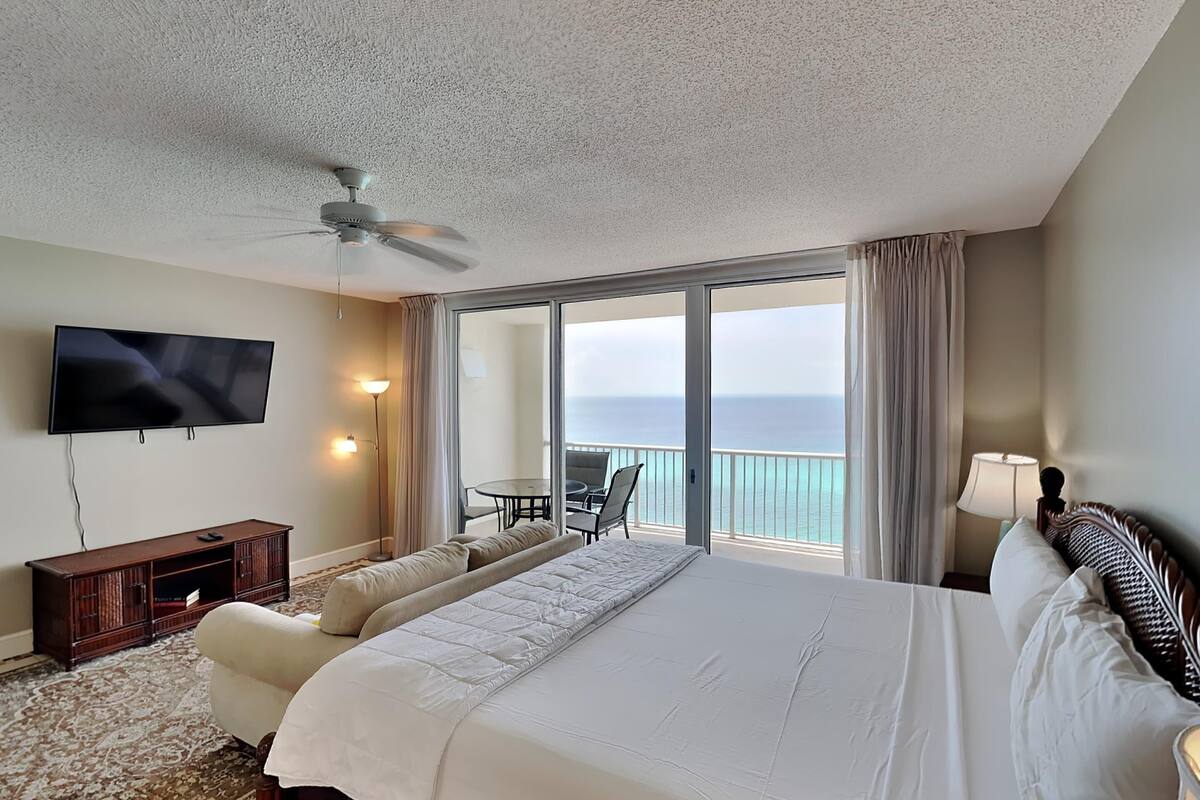A hotel room with a large bed, a television, a ceiling fan, a balcony with a view of the ocean, and a small table with chairs.