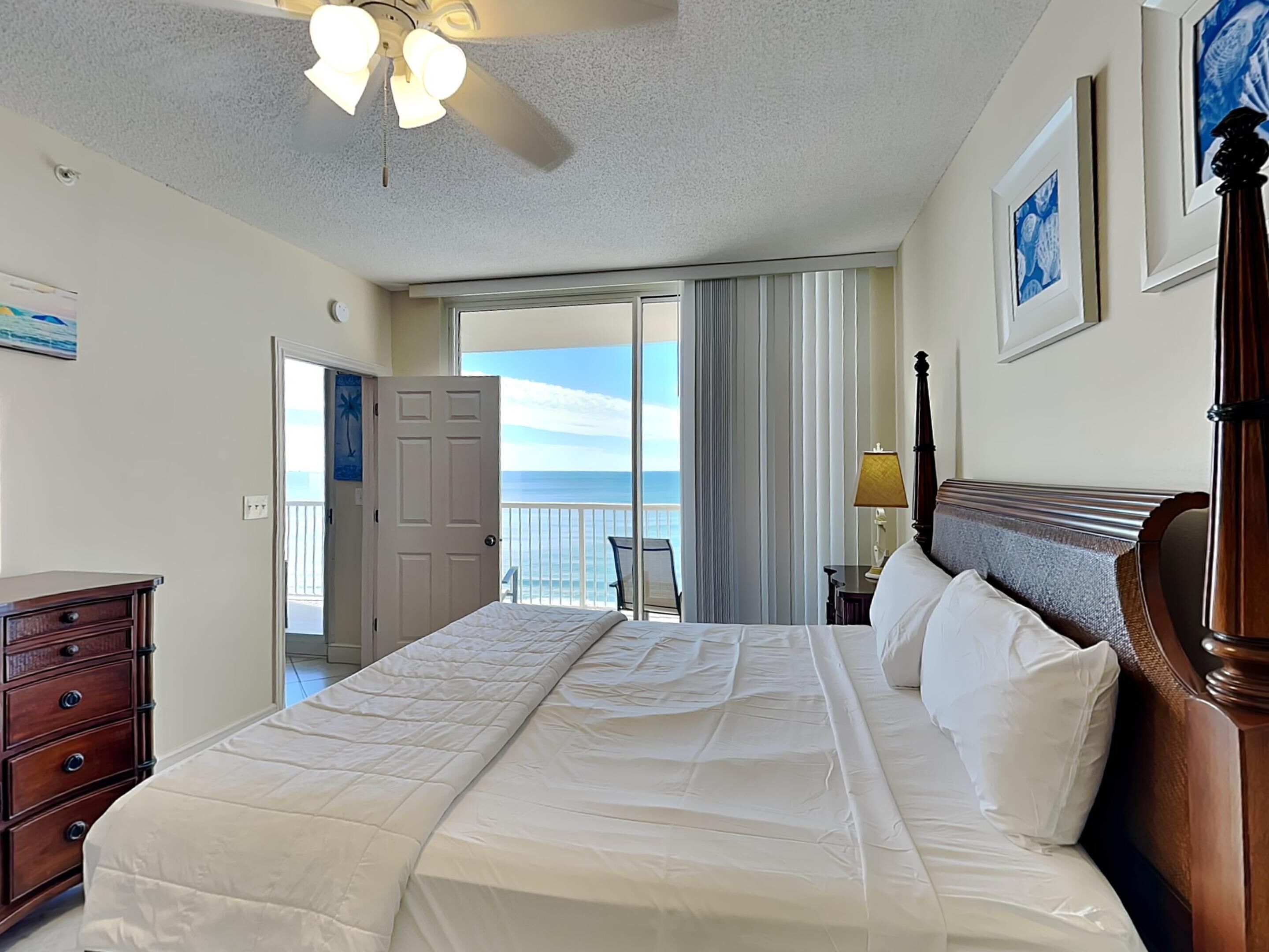 A hotel room with a large bed, a wooden dresser, a desk with a lamp, and a view of the ocean through a sliding door.