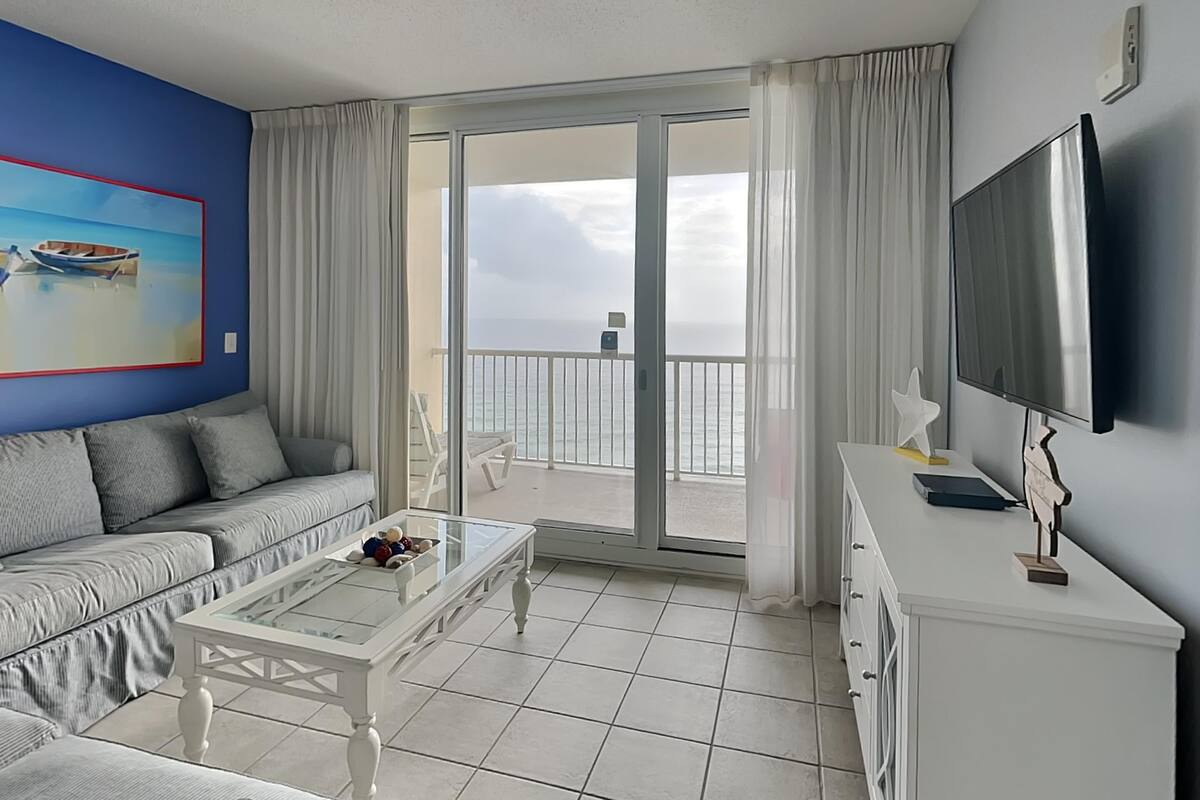 A modern living room with a sofa, a glass coffee table, a flat-screen TV, and a balcony with a view of the sea.