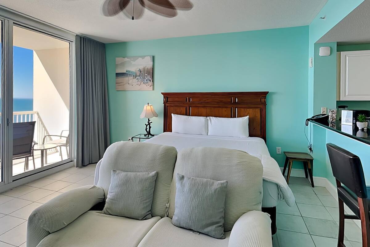 A hotel room with a bed, a desk, a chair, a ceiling fan, and a view of the balcony.