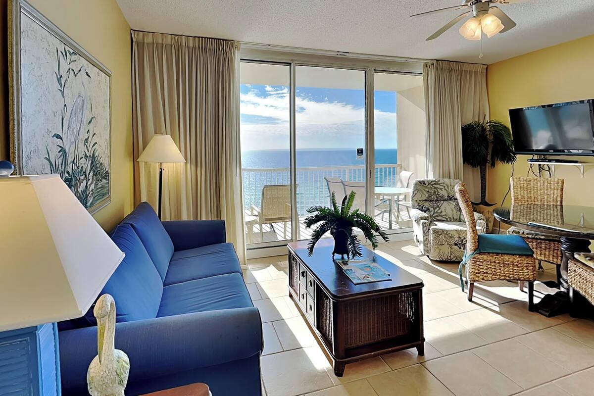 A living room with a blue sofa, a coffee table, and a TV. There is a balcony with a view of the ocean.