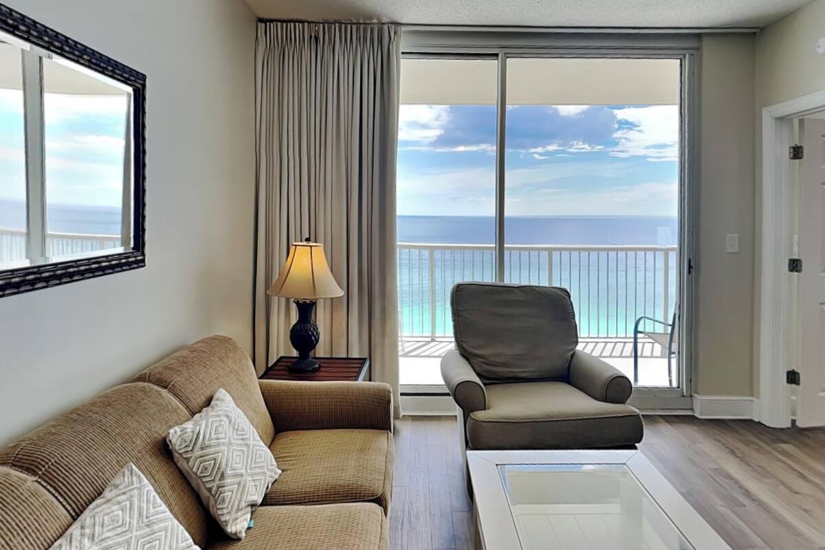 A room with a sofa, a chair, a lamp, and a view of the ocean through a large window.