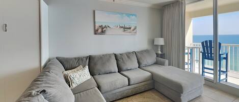 Una sala moderna con un sofá seccional gris, un tapete redondo tejido, un ventilador de techo, un cuadro de una playa y un balcón con sillas azules.