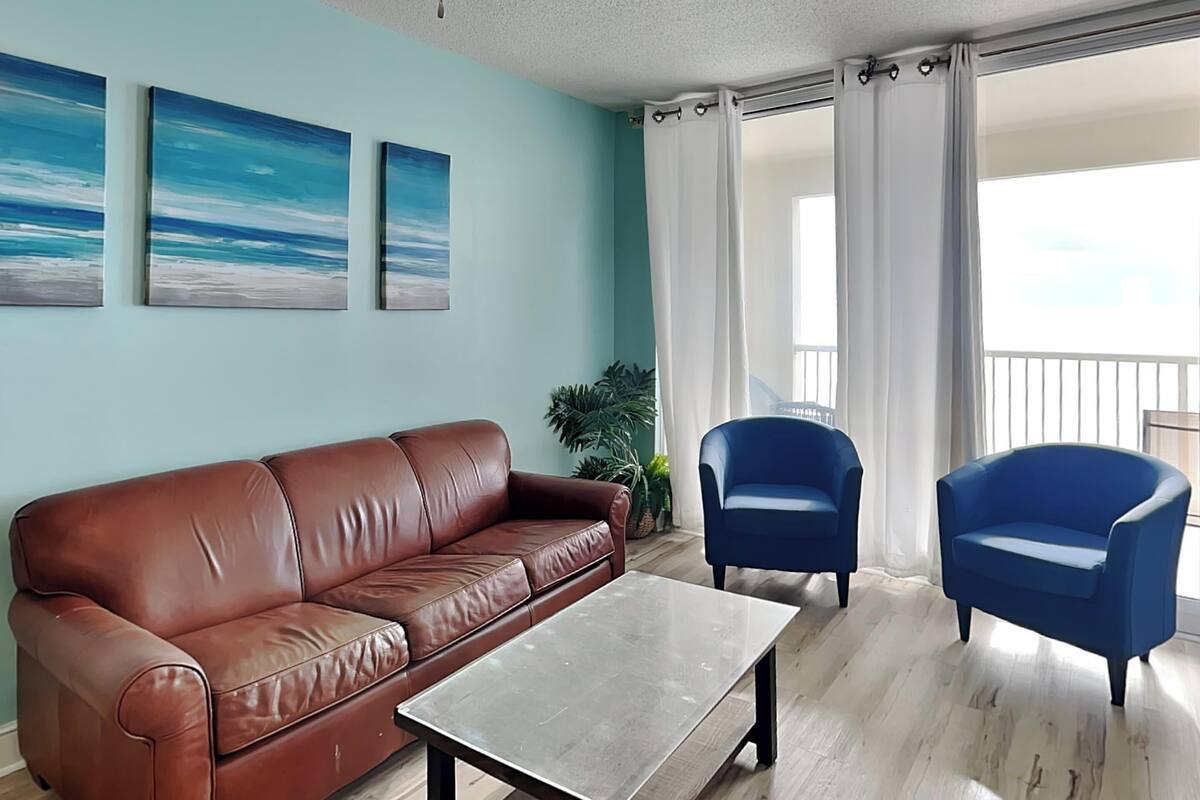 A living room with a brown leather sofa, a blue armchair, and a coffee table. There are three framed pictures on the wall and a plant on the floor.