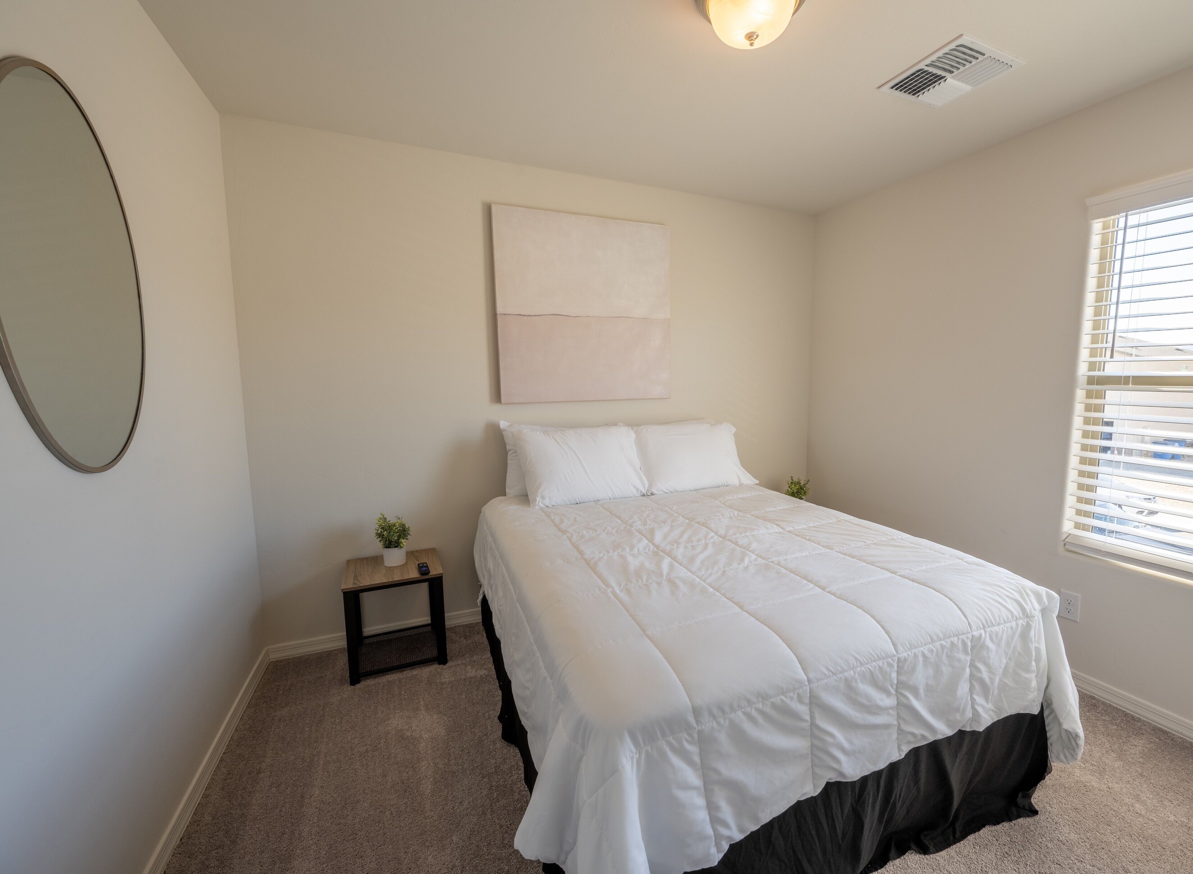 A bedroom with a bed, a small bedside table with a plant, a wall-mounted artwork, and a window with blinds.