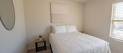 A bedroom with a bed, a small bedside table with a plant, a wall-mounted artwork, and a window with blinds.