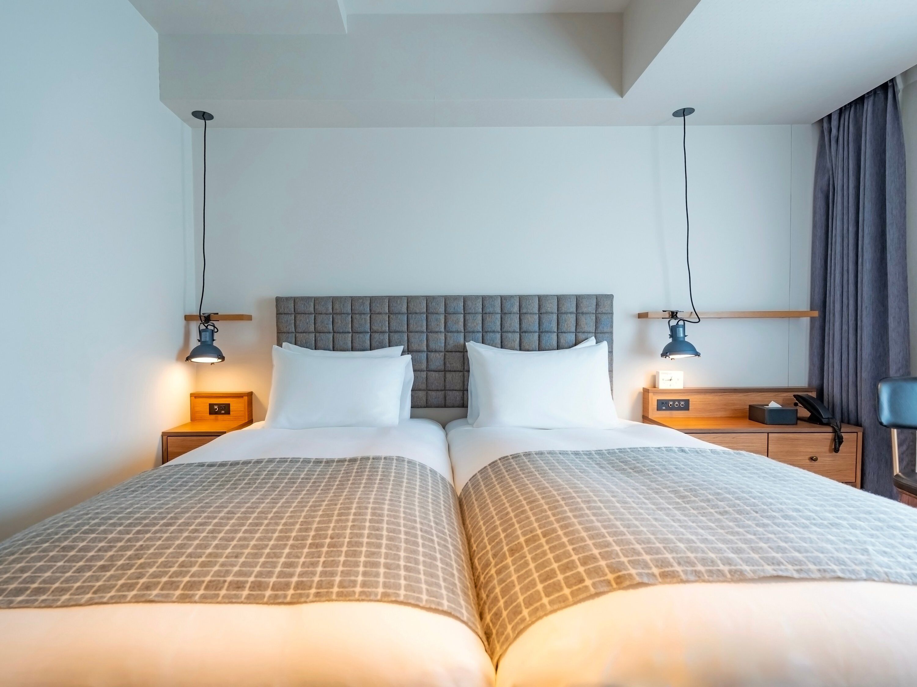 Un lit double avec une tête de lit capitonnée, deux tables de chevet avec des lampes, une horloge de chevet et un rideau sur le côté droit.