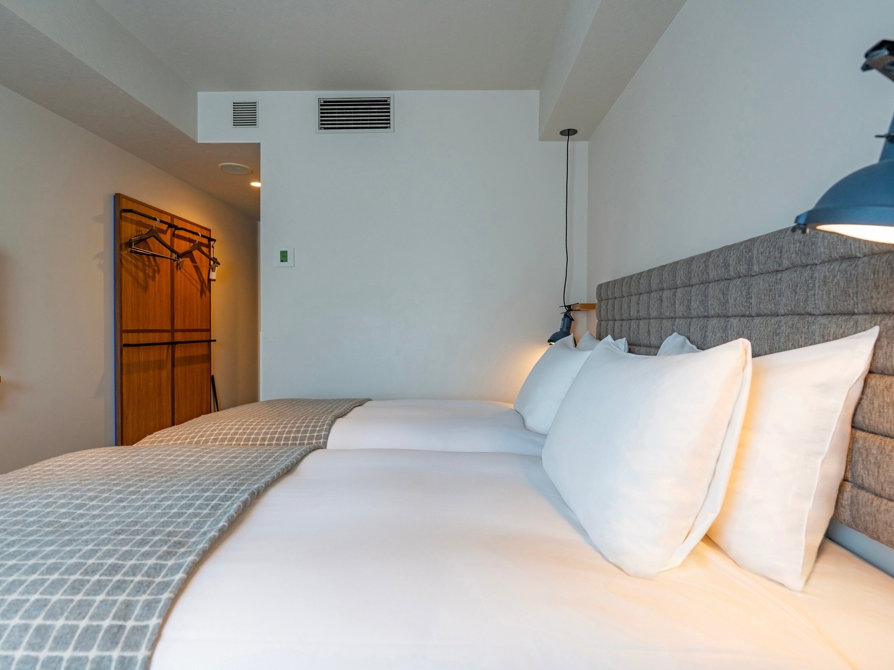 A hotel room with a bed, a wardrobe, a wall-mounted light, and a ceiling vent.