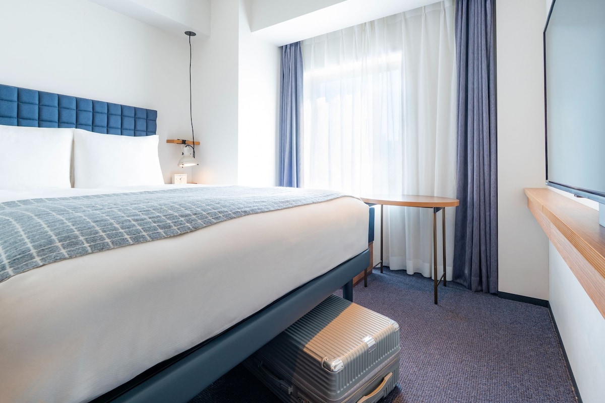 Una habitación de hotel con cama, mesita de noche, lámpara y ventana con cortinas.