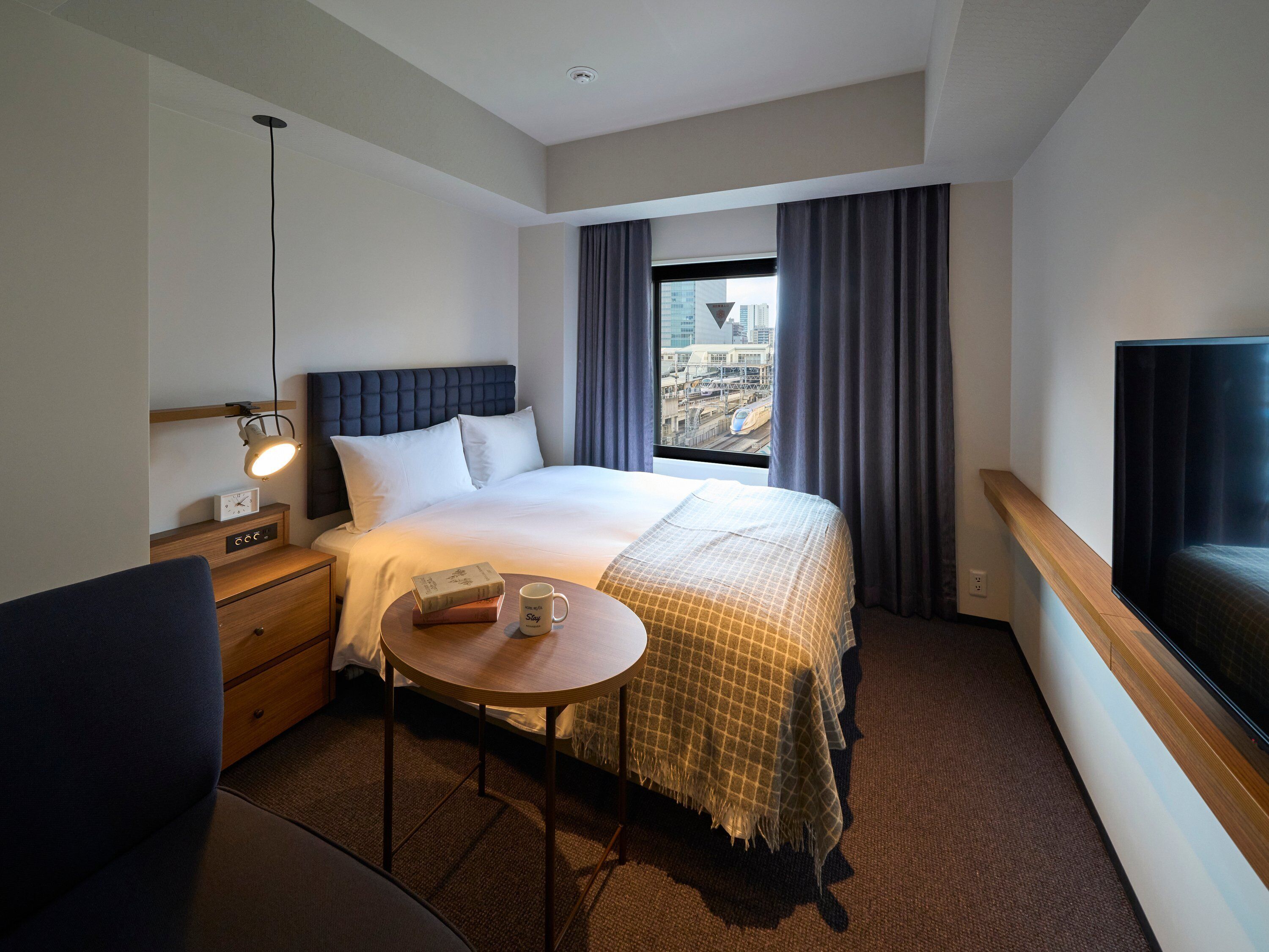 Habitación de hotel con cama, mesita de noche, lámpara y vistas a la ciudad.