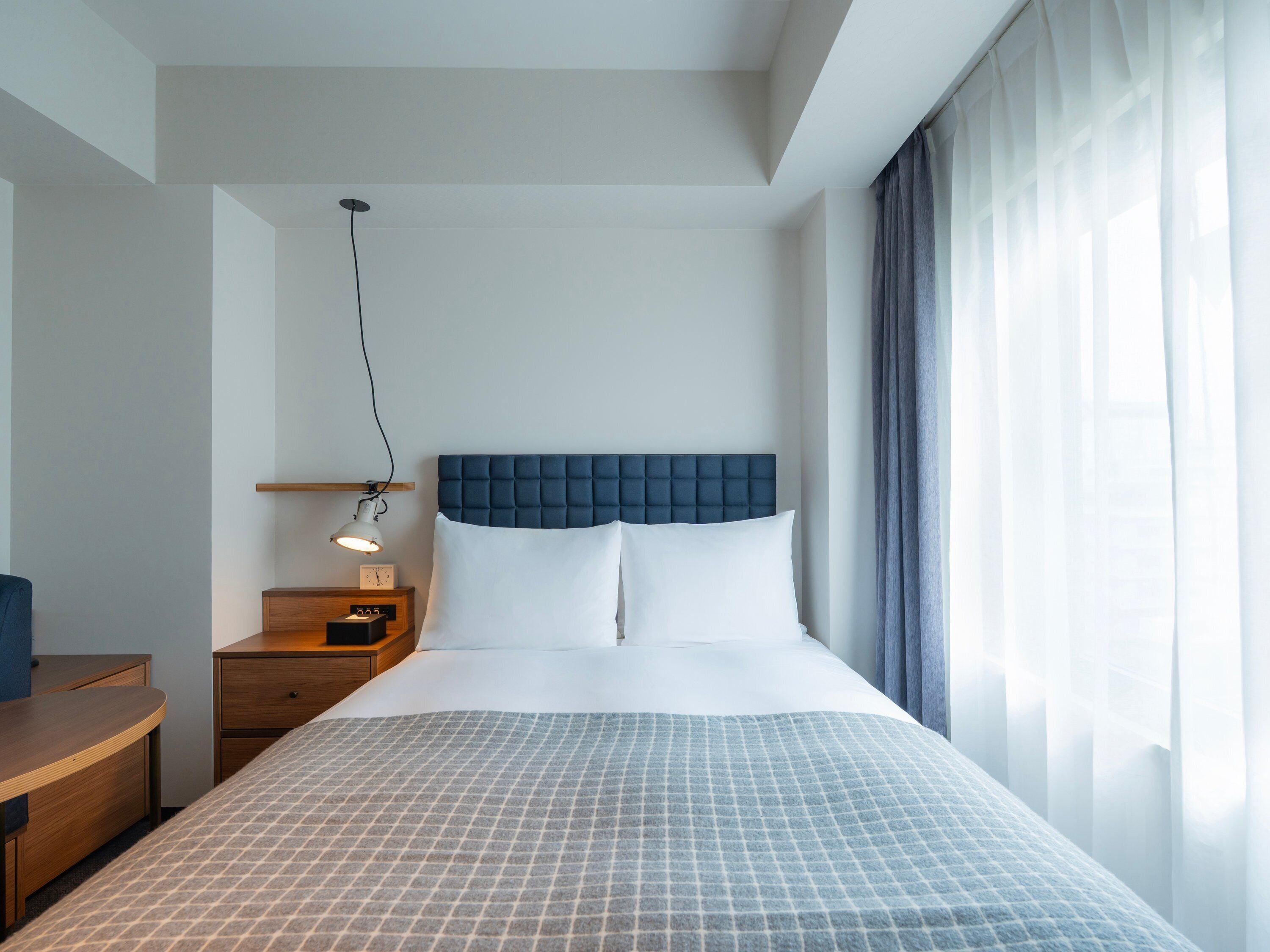Ein Hotelzimmer mit Bett, Nachttisch, Lampe und einem fensterbepflanzten Fenster.
