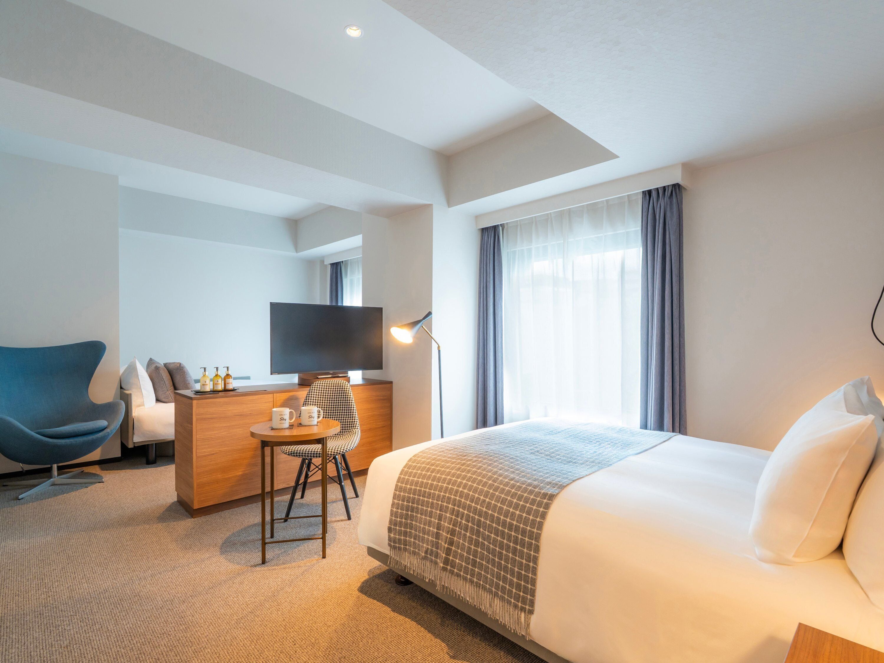 A hotel room with a bed, a desk with a chair, a television, and a window with curtains.