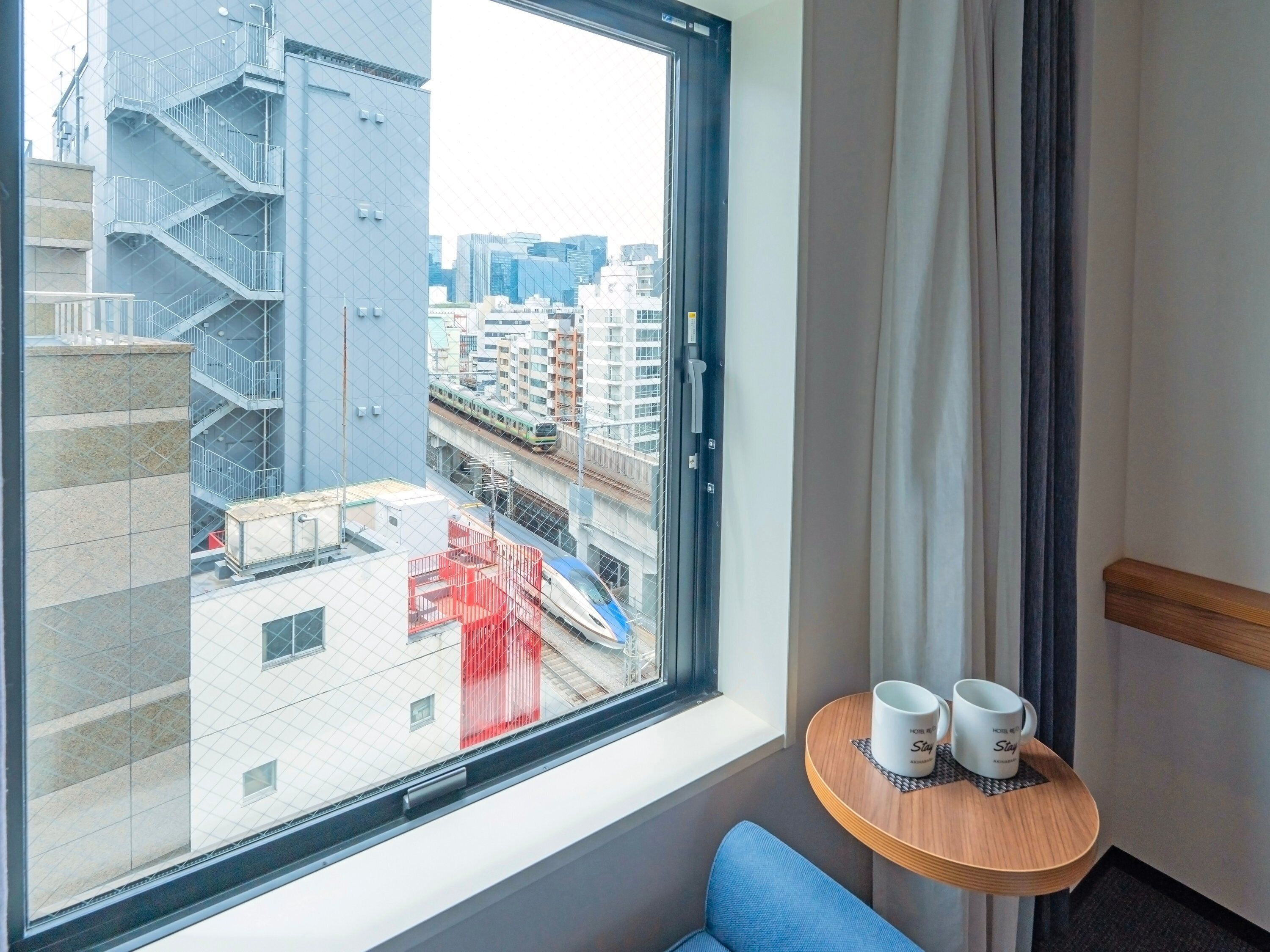 A hotel room with a large window overlooking a cityscape, a small table with two mugs, and a blue chair.
