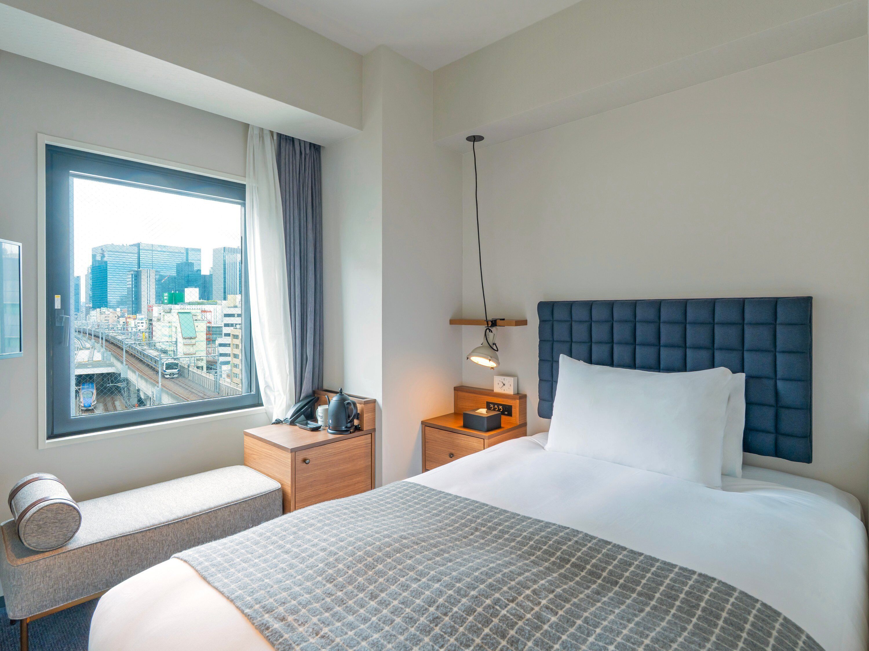 A hotel room with a bed, a bedside table, a lamp, a window with a city view, and a small seating area.