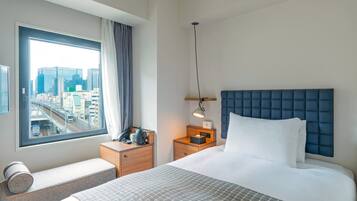 A hotel room with a bed, a bedside table, a lamp, a window with a city view, and a small seating area.
