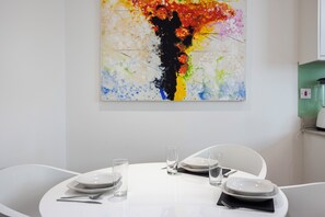 Ein Essbereich mit einem weißen Tisch, gedeckt für zwei Personen, einem farbenfrohen abstrakten Gemälde an der Wand und einem grünen Gerät im Hintergrund.