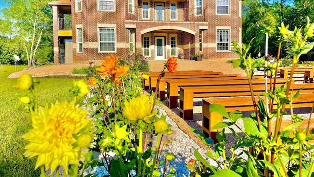 A two-story brick building with a central entrance, surrounded by a garden with benches and flowering plants.