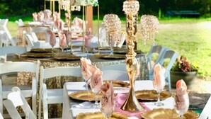 An outdoor dining area with white chairs and tables, each set with gold and pink tableware, and a central candelabra.