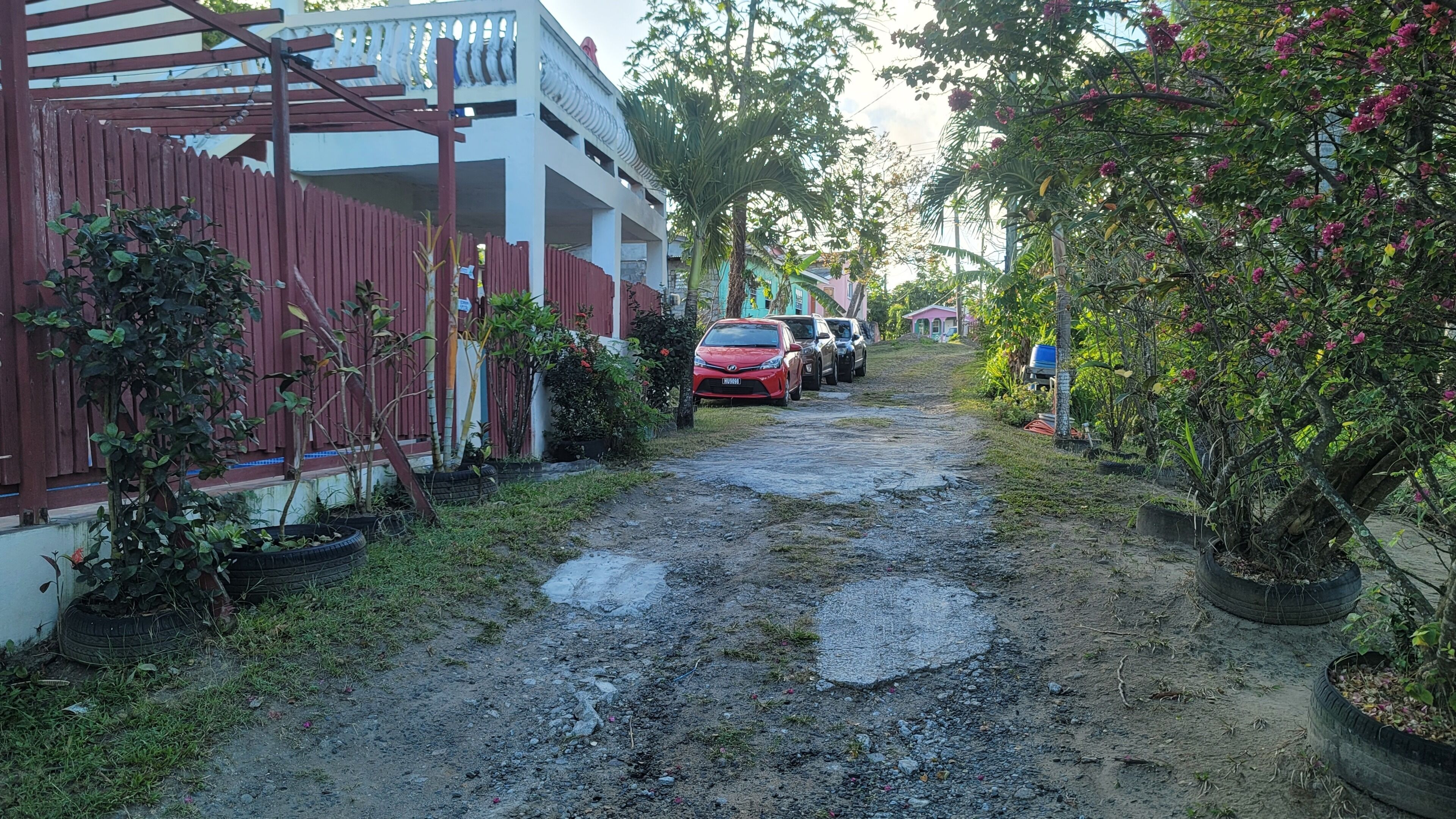 En smal, støvet sti, der fører hen til en række parkerede biler, med et rødt hegn og plantekasser langs siderne.