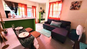 A living room with a sofa, a dining table set with plates and glasses, a fan, and a kitchen area with a window and curtains.